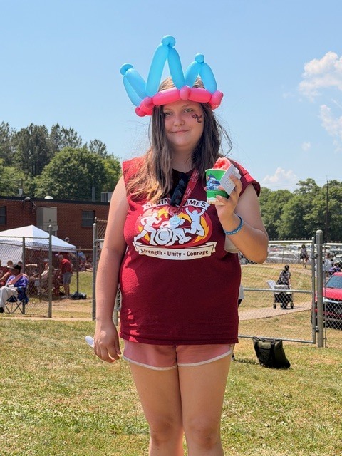 Woman wearing a balloon hat, a red shirt, and shorts standing in a grassy area with a fence.