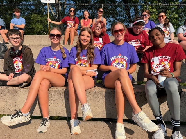 Several young people in red shirts sit on bleachers at a sporting event. They wear sunglasses and sneakers.