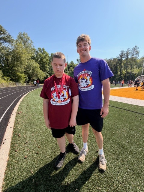 Two boys stand on a green field with artificial turf. One wears a maroon shirt, and the other wears a purple shirt. Behind them is a grassy area with trees.