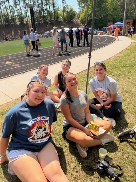 Four women sit on a grassy area; one holds a plate, another wears white shorts. Behind, a sidewalk with people and a track field.