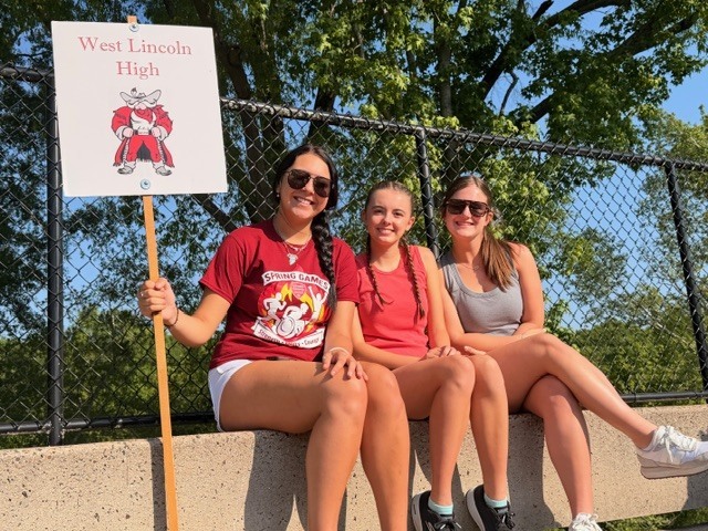Three women sit on a concrete ledge. One holds a sign for West Lincoln High. A fence and trees are in the background.