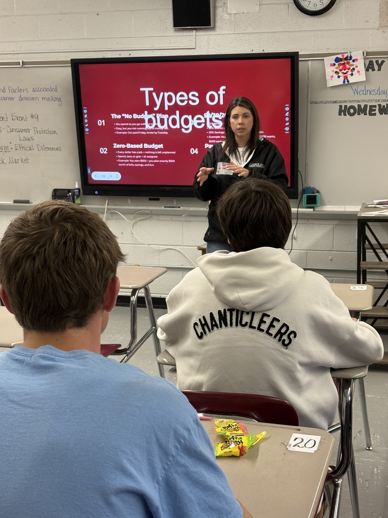 A teacher in a black jacket stands before a large screen that says "Types of Budgets". Two students, one in a blue shirt and one in a white hoodie, sit in front.
