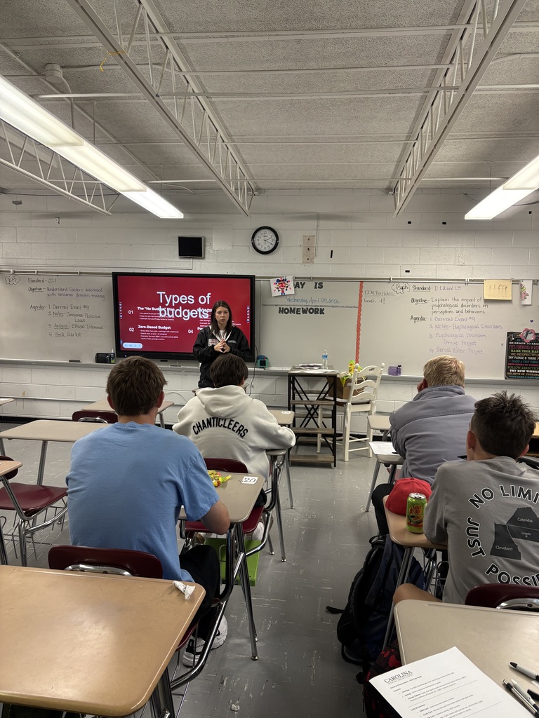 A classroom with students seated at desks facing a teacher standing in front of a screen.