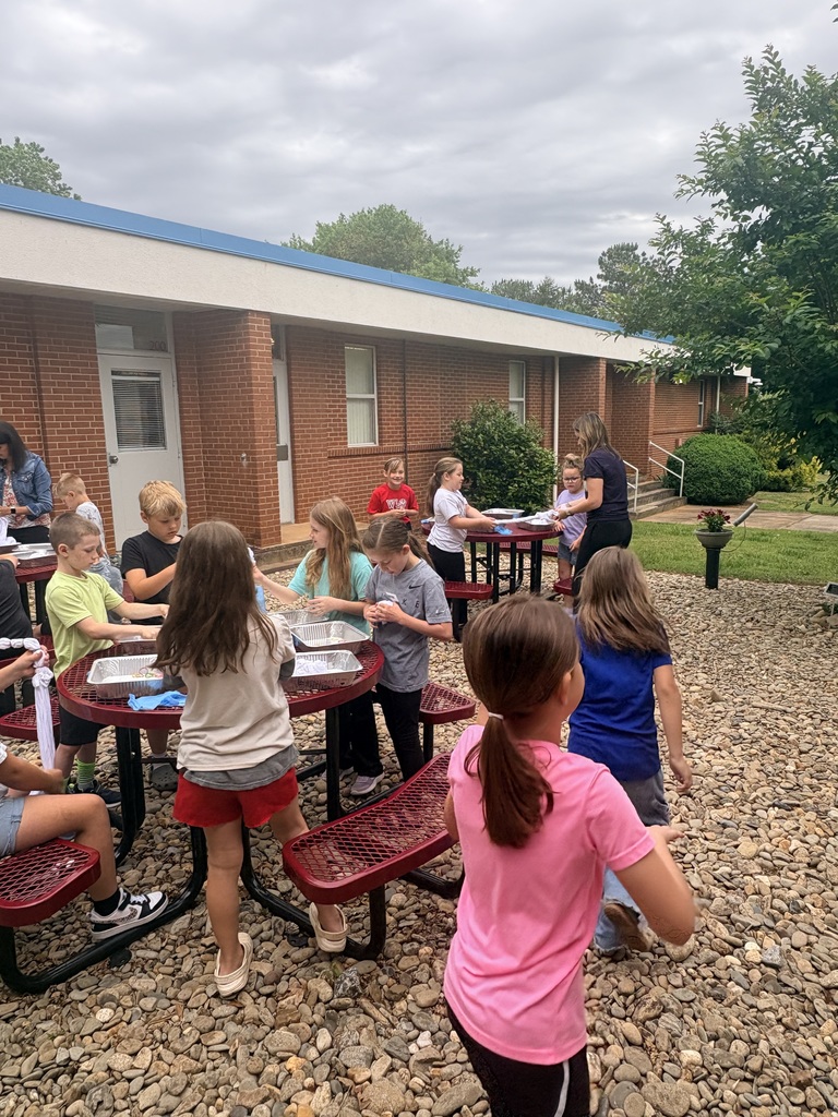 Children sit at tables in a courtyard, with a brick building in the background. Some children are seated while others stand.