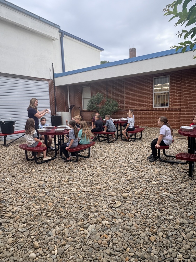 Children seated at picnic tables outdoors, with a teacher standing in front of them. Behind them is a brick building with windows.
