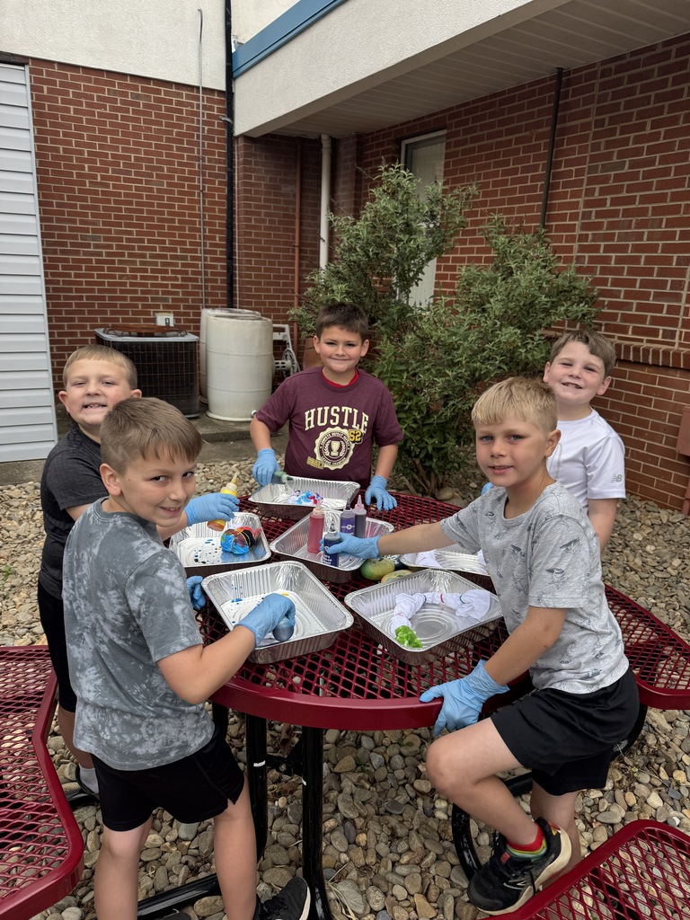 Five boys wearing gloves sit around a red table. One holds a tray. A brick building is behind them.