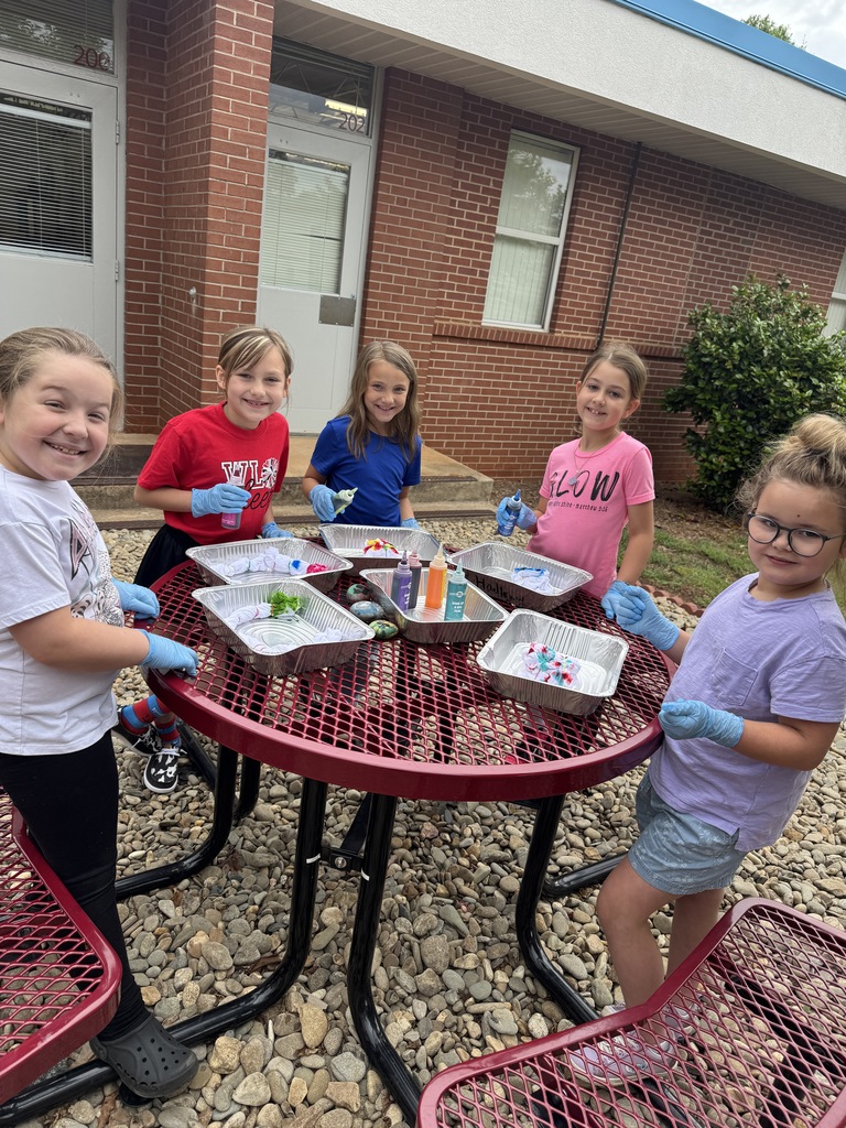 Six children in protective gloves gather around a table filled with colorful art supplies, standing outside a brick building.