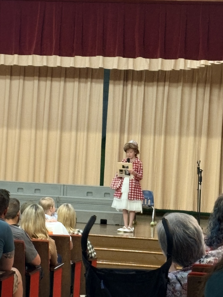A young girl stands on a stage with a book, performing for an audience seated in rows.