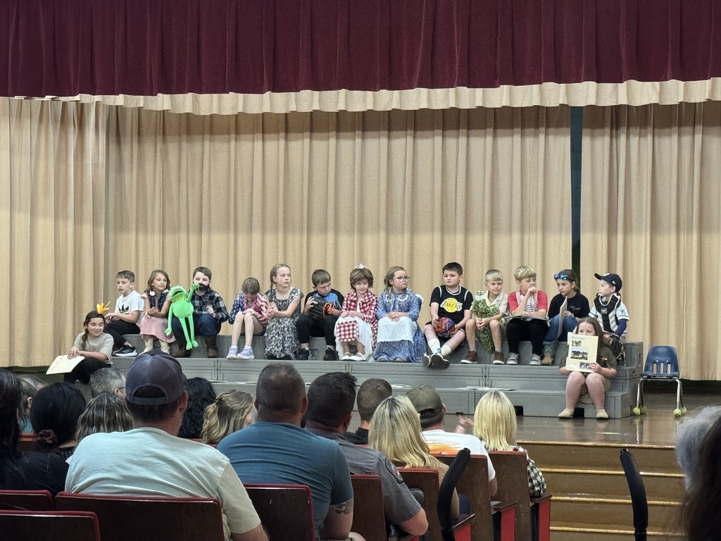 A group of young children stand on a stage holding props. Some have costumes, others wear casual clothes. Audience members sit in chairs facing the stage.