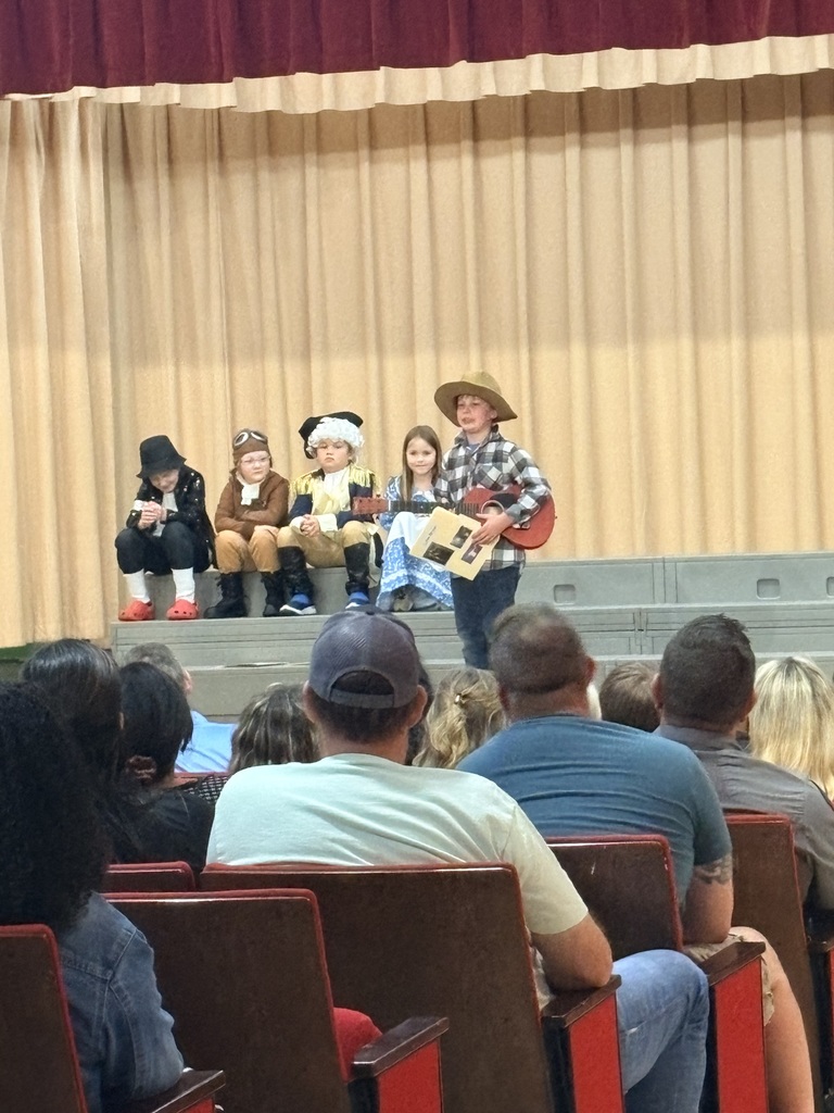 A group of children in costumes perform on a stage while an audience watches. The stage has a backdrop.