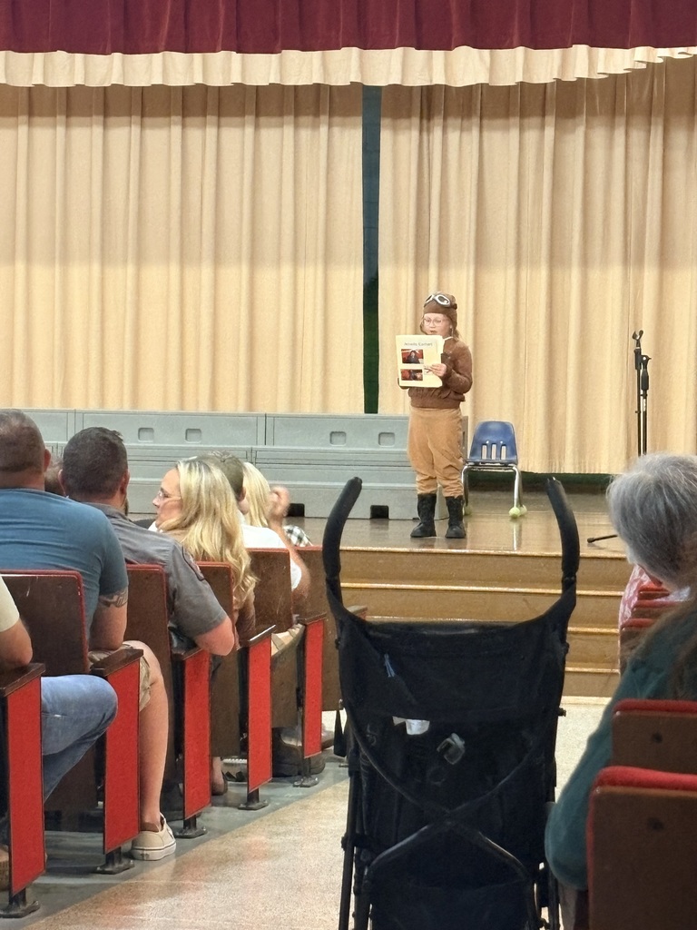 A person stands on a stage with a book, wearing boots and a costume. Several people sit in chairs, watching. The stage has a curtain backdrop.