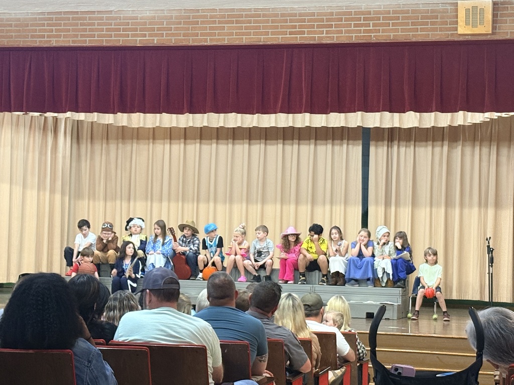 Many children dressed in costumes sit on stage, with a large audience seated in chairs in front of them.