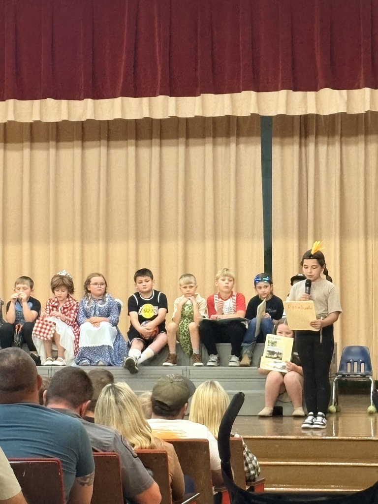 A group of children stand on a stage, with one child holding a book. They are seated on the floor, and the stage has a curtain backdrop.