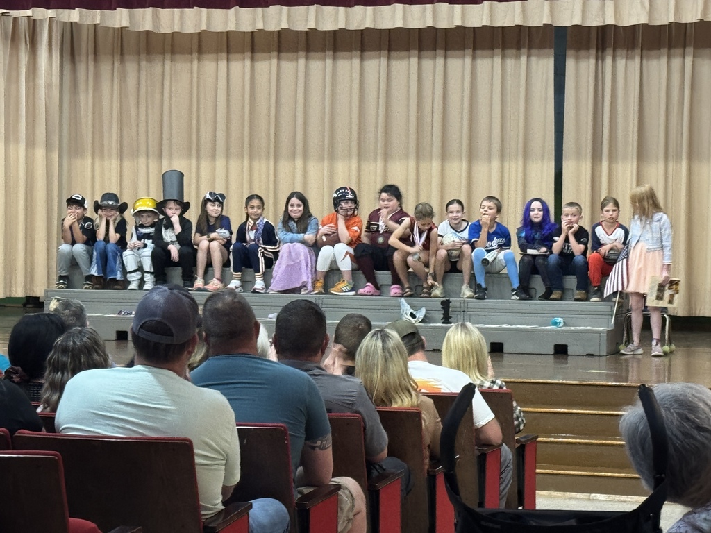 An audience watches as a group of children, dressed in costumes, stands on a stage with a brown curtain backdrop.