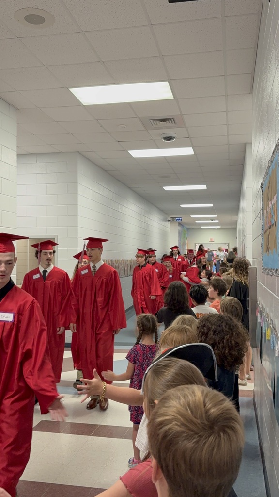 Graduates wearing red gowns walk through a hallway while people stand along the sides to watch.