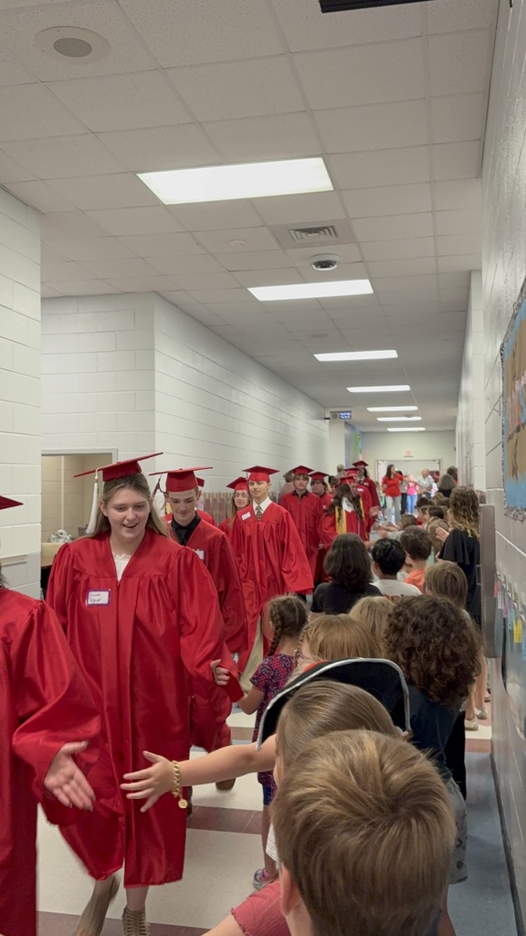 Students in red gowns and hats walk down a hallway as others watch. White walls, ceiling lights, and bulletin board visible.