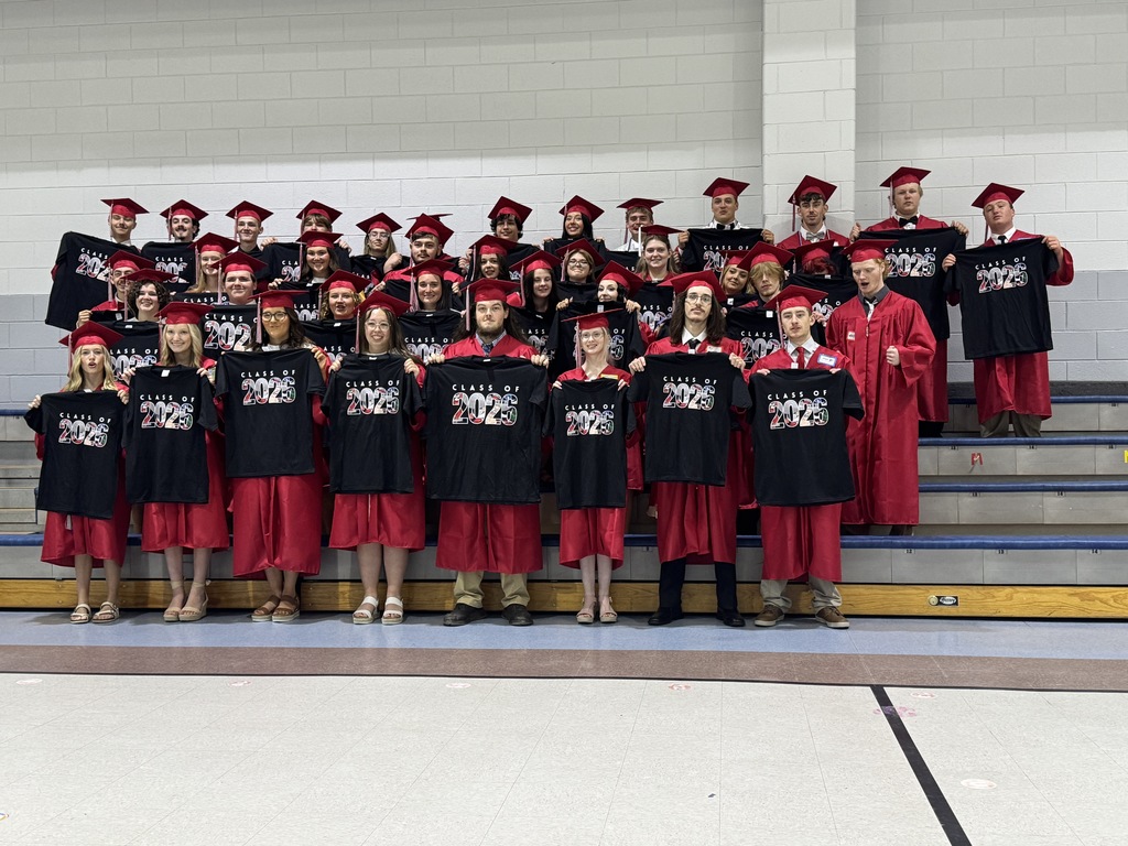 A group of people in red graduation gowns and caps holding black T-shirts pose on a stage.