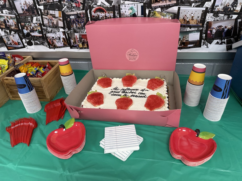 A pink box containing a cake with strawberry icing sits on a green table. Plates, cups, and napkins are nearby.
