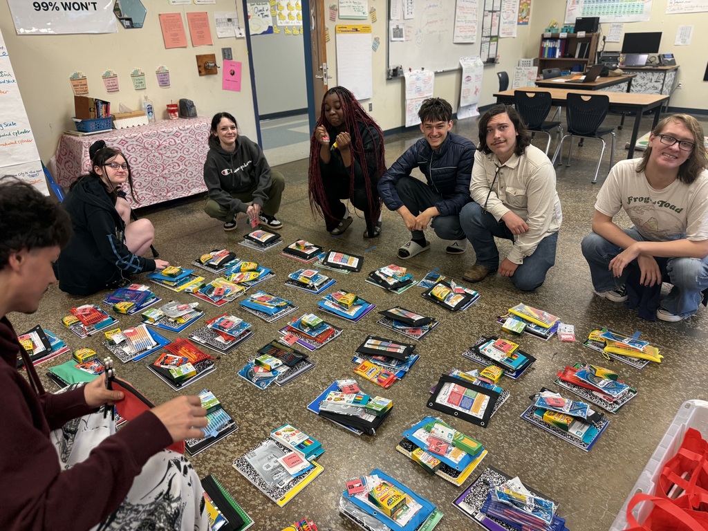 A group of people are gathered around a table filled with various items, including books and art supplies.