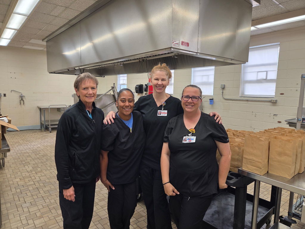 Four women in black uniforms stand in a kitchen, posing for a picture. A large hood vent is above them.