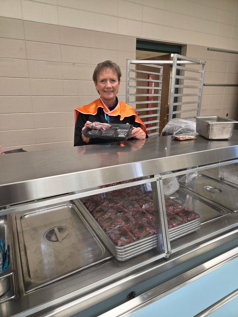 A woman in a reflective orange vest holds a black tray in a kitchen area with a blue counter and stainless steel shelves.