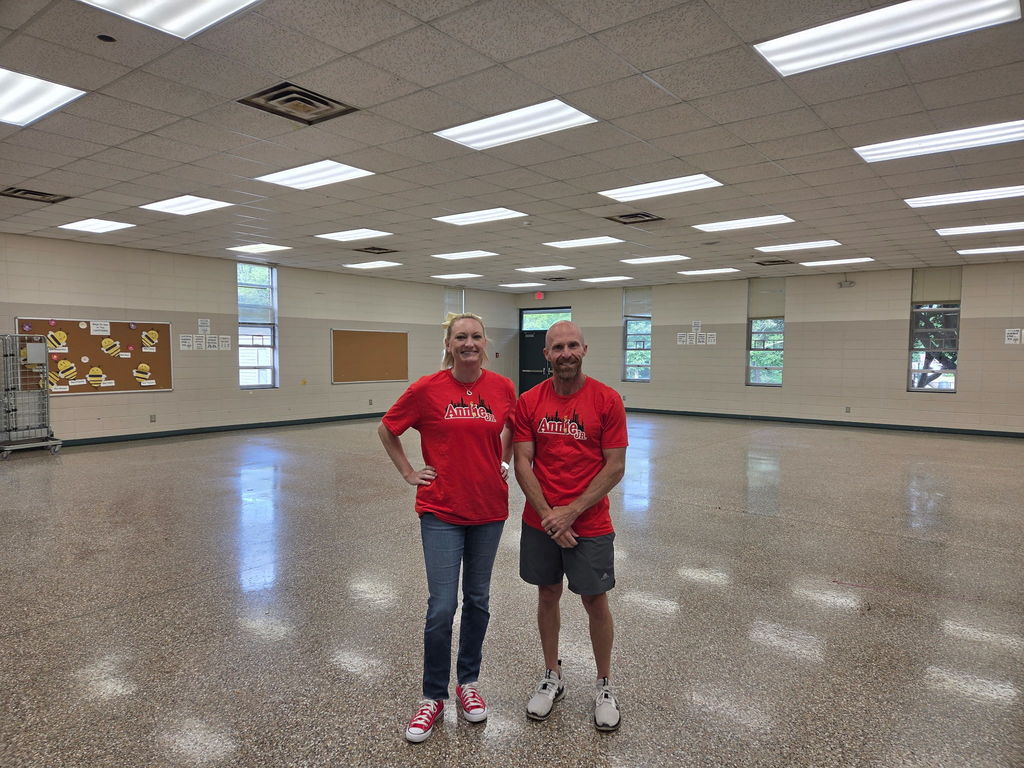 Two people in red shirts stand in a room with windows and ceiling lights. The floor is shiny.