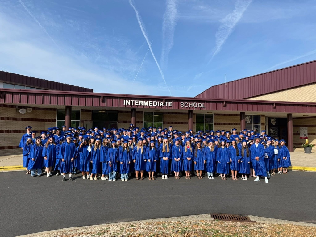 A group of students in blue gowns stands in front of a school building with "INTERMEDIATE SCHOOL" on it.