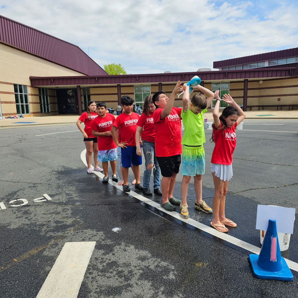Group of children in matching shirts lined up in a wet road, holding water bottles, with a building in the background.
