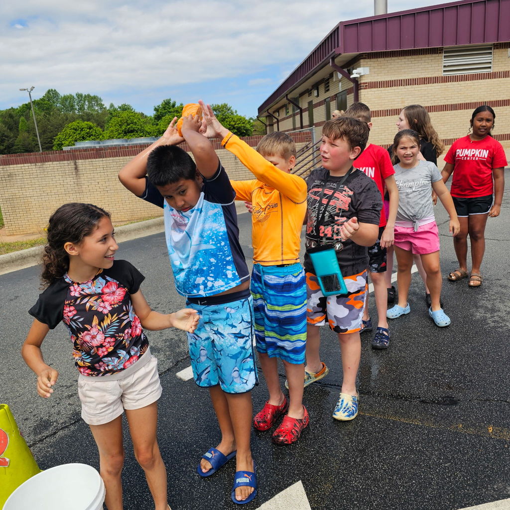 Kids in swim clothes and sandals stand in a parking lot, one boy holds hands with another.