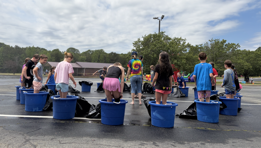 People standing in blue buckets filled with water, gathered in a parking lot with a cloudy sky.