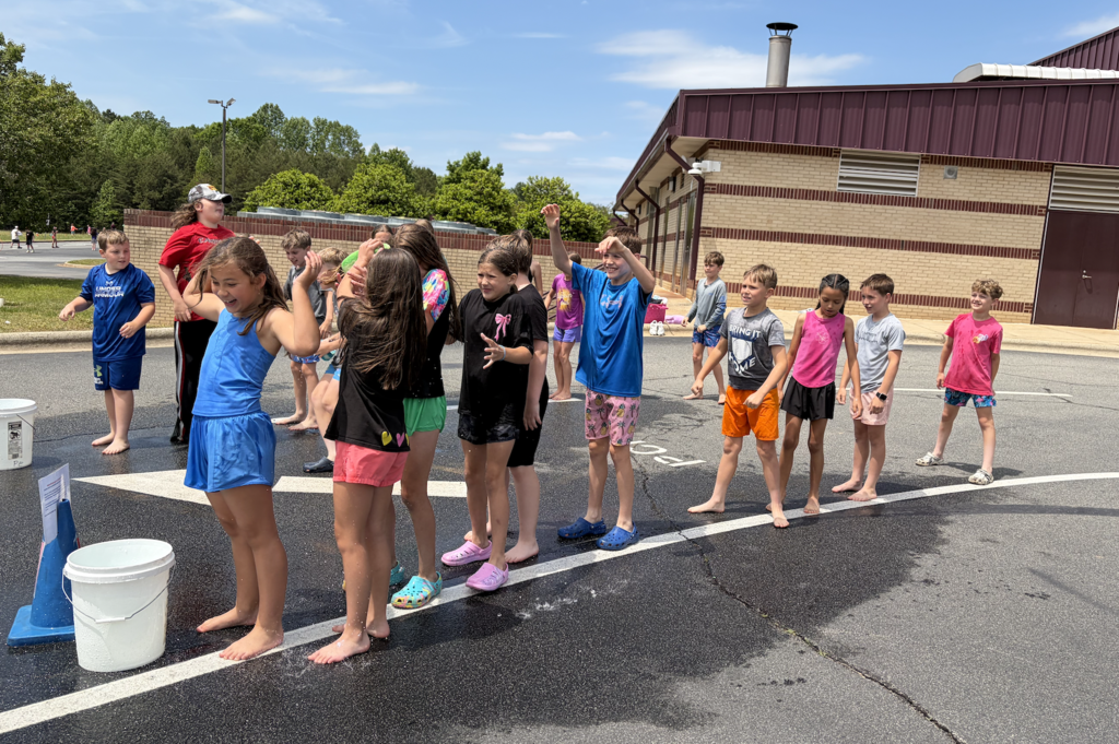 Several children wearing bathing suits stand in a row in a parking lot with a building in the background.