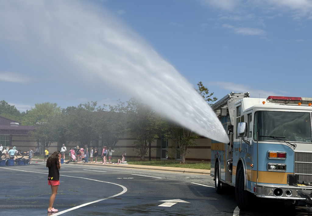 Water spraying from a fire truck nozzle. A person stands in the wet road. Background shows a building and trees.