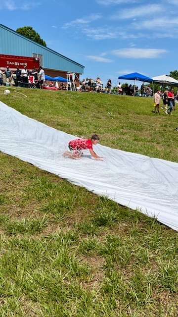 Child sliding down a long, white slip and slide on a grassy field. Behind them is a building and several tents.