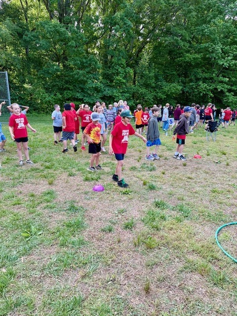 People in red shirts stand in a field with grass, some holding frisbees. Trees are in the background.