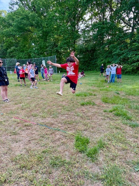 A person in a red T-shirt jumps in the air on a grassy field. Many children and adults are behind them.