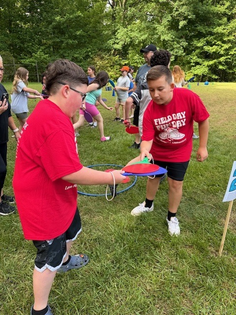 Two kids in red shirts play with paddles in a grassy field, with others engaged in various activities.