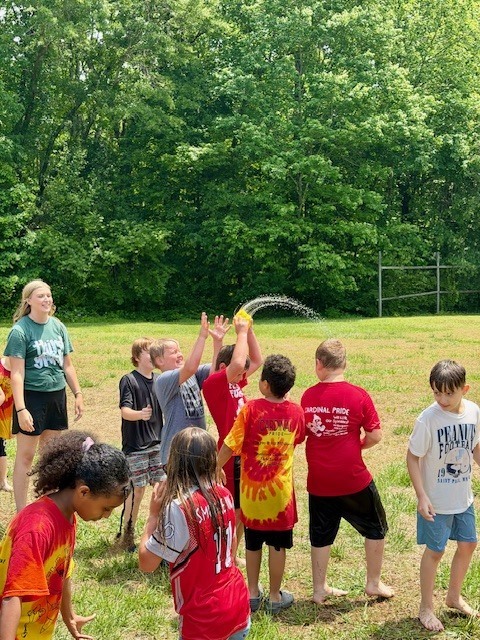 Kids standing in a grassy field, one holding a water balloon. Green trees in the background.