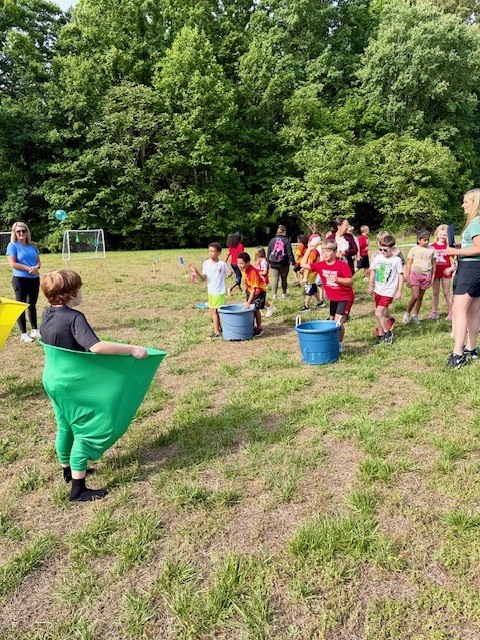 A group of people, including children, play outside on a field with buckets and water balloons.