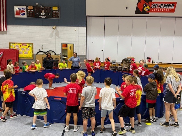 A large group of elementary children in matching shirts gathered around a blue plastic fence like structure, in a school gym, playing and observing their classmates playing Gaga ball.