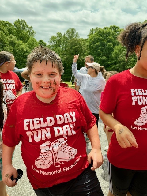 A boy with mud streaks on his face walking through a crowd in a red Field Day shirt. Behind him, another person in a similar shirt.