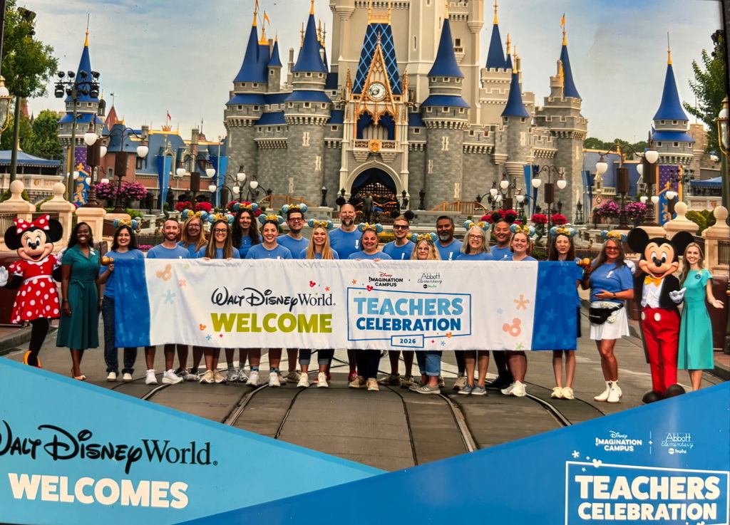 A group of people in blue shirts stand before a banner outside a castle. They are joined by Mickey and Minnie Mouse.