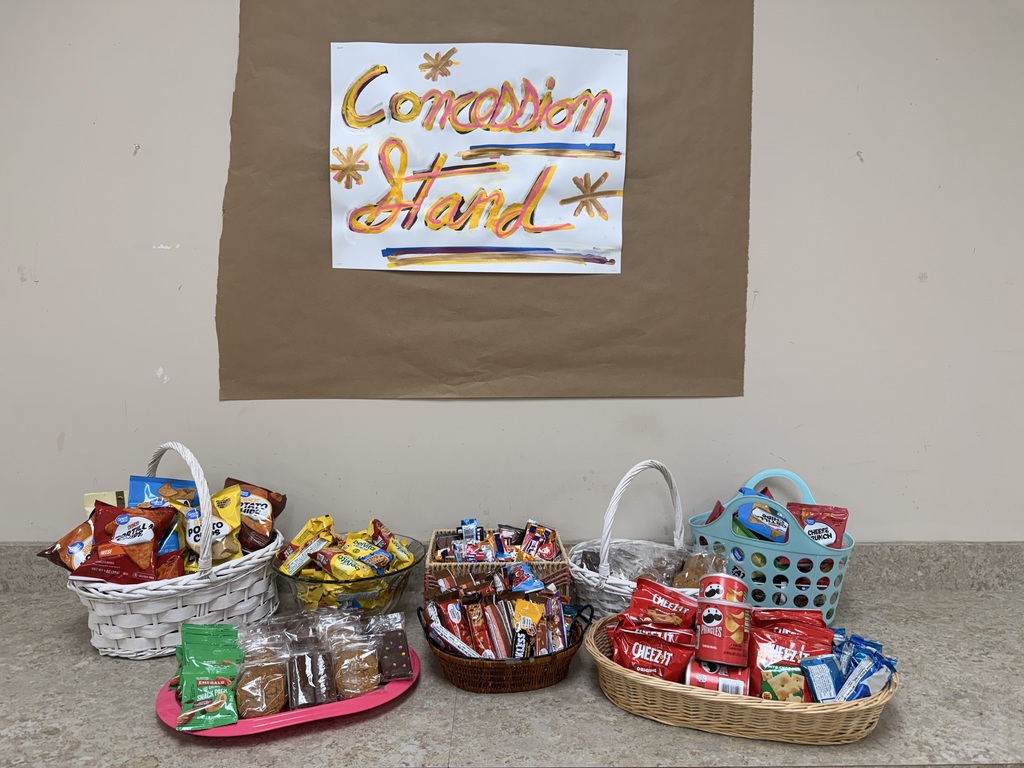 A concession stand display featuring several baskets and bowls filled with assorted snacks. A white handmade sign with "Concession Stand" written in yellow and pink stylized lettering with gold accents is taped to a brown paper background on the wall above. The snacks include bags of Doritos, Lay's potato chips, Cheez-It crackers, Pringles cans, packages of cookies, brownies, Rice Krispies Treats, and various candy bars. The items are organized into white wicker baskets, a glass bowl, a brown woven basket, a blue plastic basket, and a pink tray on a neutral-toned countertop.