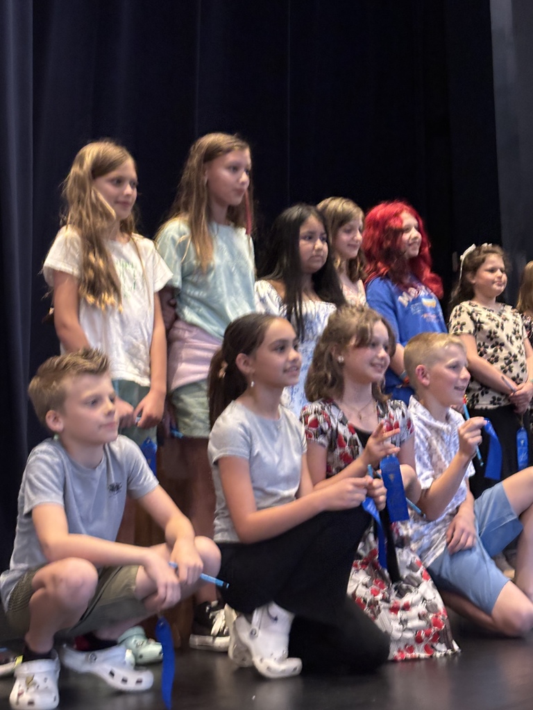 Group of children wearing casual clothing with varying hair colors, standing and sitting on stage. Blue ribbons are visible on their clothing.