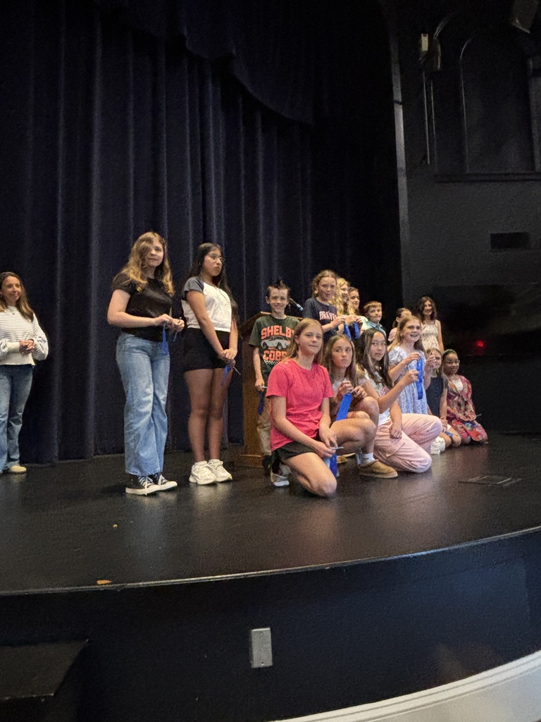 A group of young people on a stage, some standing and some kneeling, under a blue curtain.