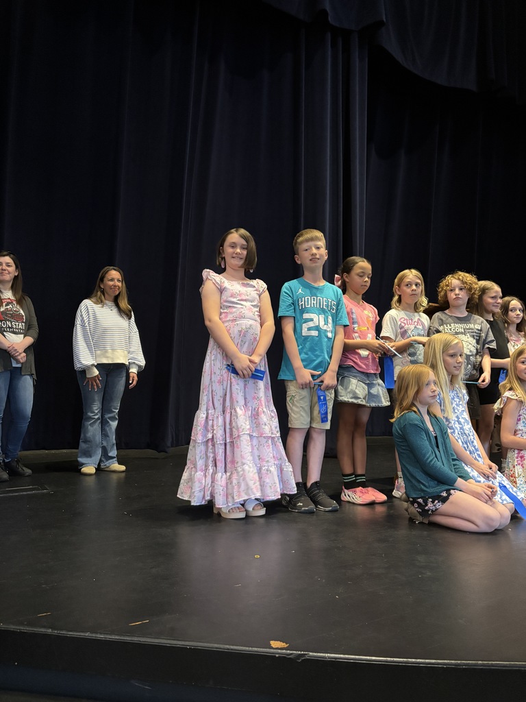 Kids on a stage, some standing and others sitting, in various outfits, with a black curtain in the background.