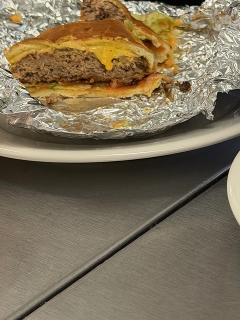 A burger with cheese on a foil-covered plate sits on a white plate on a metallic surface.