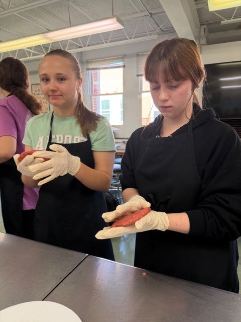 Two women in aprons and gloves prepare food at a kitchen table, with sunlight coming through windows.
