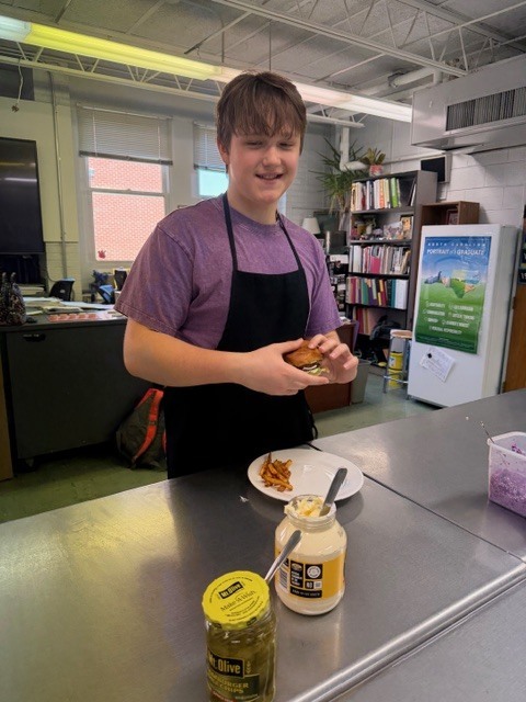 A person in a purple shirt and black apron holds a sandwich near condiments and a plate.