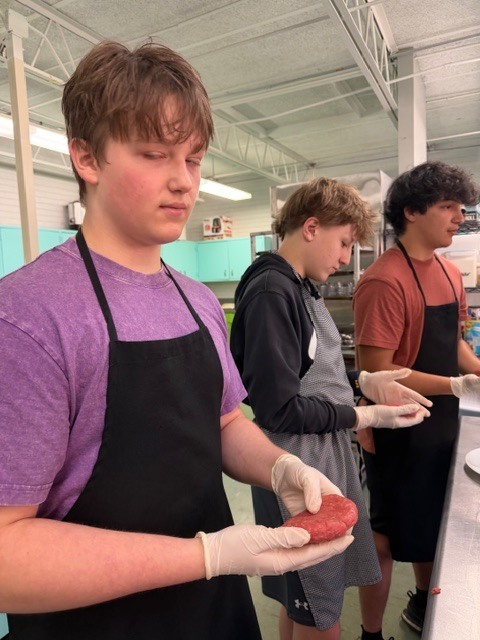 Three people in a kitchen. The person in the front wears a purple shirt and black apron. The others wear hoodies and black aprons.
