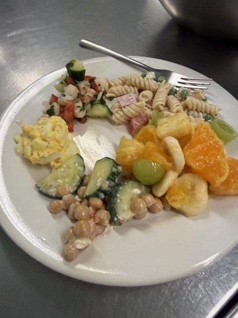 A white plate with assorted foods: eggs, pasta, beans, vegetables, and fruit, placed on a silver table.
